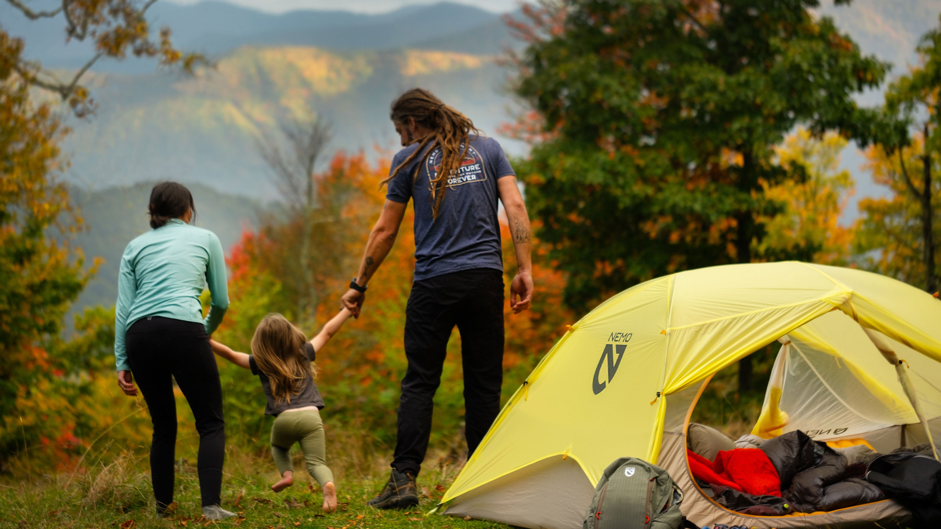 An adult and a child holding hands stand beside a yellow tent in a scenic outdoor setting with autumn foliage in the background. A backpack is placed near the tent.